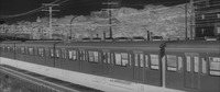 Again in negative, rightwards pan captures the bullet train entering the frame from the Left background and moving in the direction of Right foreground, captured at an angle (on the train’s left side) and stretching across the entire frame as does the rail bridge underneath it, the electrical powers and tower lines, and a line of trees seen above, all of them at an angle parallel to the train.