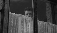 A man peers through a window over a lace curtain.