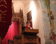 Image of a reverend speaking at a podium in a church. There is a red curtain behind her and frescoed walls along the right side of the image.