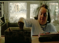 Color screenshot from a fiction film showing one female adult with Down syndrome in close up on top of a ladder in a small family store. Her hair is being blown about to simulate the effect of a bomb explosion nearby.