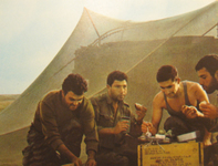 Color candid photo of 4 uniformed Mizrahi IDF armored corps men taking a break from battle to eat and drink together in the desert.