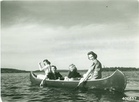 A black and white photograph of four women in a canoe on a lake. They are all looking at the camera and smiling as two of the women push their oars through the lake water, and the other two sit in the middle of the canoe.
