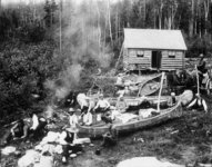 A group of scattered forest rangers, sitting, socializing, and relaxing on the grass alongside canoes. A cabin is in the background.