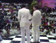 Color still of two men (Two Beat) performing on stage, seen from perspective of stage. Men’s backs are visible in foreground, and audience, faced forward, are visible in foreground.