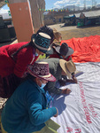 A close-­up of a group of Andean women sitting on the floor embroidering a big cloth. They are wearing their traditional hats.
