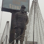 A statue of Colonel Ojukwu stands at Niger Bridge, Anambra State, Nigeria. The statue stands beneath gray skies among city billboards and pyramids of lights. Relatively understated, the statue is entirely dark gray in color and depicts Colonel Ojukwu in a Biafran army uniform and cap.