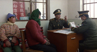 Two soldiers read paperwork and smoke, wile two people in winter clothes wait alongside the desk. Red signs with white calligraphy can be seen through the window behind them.