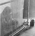 Photo of a veiled Mizrahi woman in a museum exhibit weaving on a loom next to a small stove with a kettle and a tray with a smaller kettle.