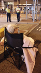 Photographed from the back, Wong Fung-Yiu (Old Lady Wong) sits in a folding chair in the street. She has white hair, wears glasses and an air pollution mask, and is covered with a blanket. Two security officers stand across the street, facing her