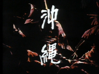 White calligraphy for "Okinawa" is superimposed over the reflection of explosions on rocks in a bay.
