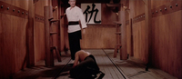 Image of a person crouching in front of another person in an empty room. In the background, there is a large white square on the wall with black calligraphy on it.