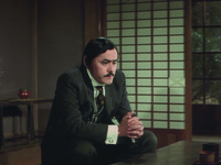 Aochi crouches in his impeccable suit, center frame, in a different part of the sitting room, his eyes slowly turning away from Sono’s nudity, his expression troubled. Background Right is a purple wall and small table with a small, elegant red vase in archaic style; on the Left is another shōji.