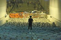 A young man stands alone with his back to the camera among rows of concrete sewage-cover road blocks. Past the barrier gates several yards in front of him people walk along a busy city street