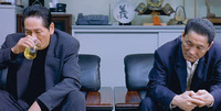 A film still of two men in suits sitting on the left and right side of the image. Between them, in the background, is a shelf full of various items, including a display plate with calligraphy on it.