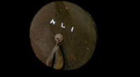 Closeup of a wooden disc, upon which the main character has chalked "Ali."