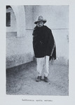 Photograph of a man standing near the corner of a room. He wears a hat, white trousers, and a dark-colored serape or shawl with a geometric design on one side. His eyes appear to be closed or partially closed.
