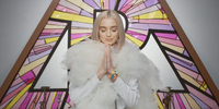 Poppy prays with her eyes closed downward, in a white tulle prayer robe in front of a pink stained-­glass P. She wears a multicolored flower and pearl bracelet and has long pink nails.