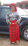 A woman stands in front of a car wearing red, black, and white aso ebi.