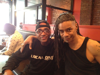 Two men sitting in a restaurant booth, arms around one another; one wears a T-shirt with the logo “Local One.”