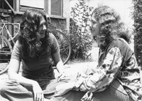 Black-and-white photo, side view. Sitting on the ground, the two look toward each other, smiling. Both have long, curly hair. A bike and a bungalow are in the background.