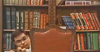 Color screenshot from a fiction film showing an adult with Down syndrome. He crouches in front of shelves full of books, with his body hidden behind a chair, only his face and hands are visible.