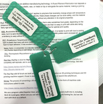 Three rectangular, green, plastic tabs are connected with a metal keychain and labeled with the printer name and settings with which they were produced.