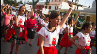 A close-­up of a group of young women in a street protest. They all are wearing traditional Andean skirts and T-­shirts with the logo of We Are 2074 and Many More. Women have one hand raised in a fist. Some of them wear over their skirts drawings of women’s reproductive organs.