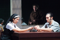 A nun and a man in handcuffs clasp their hands in prayer over a Bible as they sit at a table. A third man in monk’s robes gazes from behind a window.