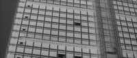The camera, at a low angle, pans vertiginously up and across the façade of a contemporary office building, a square, minimalist design of endless partitioned glass windows. The façade takes up almost the entire frame, with only a patch of sky glimpsed to the Right side of the building. The shot is entirely printed in negative (i.e., blacks and whites are reversed).