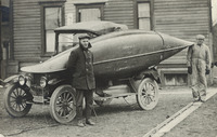 A photograph of a canoe being transported by car. The canoe is strapped horizontally on the side of the automobile, covering the doors.