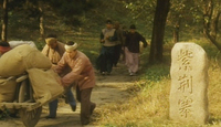 Image of a group of people following another group of people pushing a wagon down a path. In the right half of the image, there is a larg tombstone engraved with calligraphy.