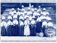 Butler directs twenty-four musicians. All wear tall, triangular hats, white blouses, and long, dark skirts. Each holds her instrument. One has a large drum. The band name is written on the drum head.