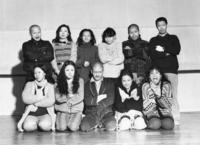 This is a black and white cast shot showing eleven people, approximately in their thirties, arranged in a studio facing the camera in two rows. Five people kneel in the front row and six stand behind them. All eleven have their arms crossed or hands crossed in front of their torsos, and each makes a different strange facial expression, such as smirking, grinning, looks of exaggerated shock or suspicion, or unsettling stares. One woman sticks out her tongue. Three of the men have shaved heads, and all of the women have long, often curly or wavy black hair. They are bundled in layers of everyday winter clothing, including sweaters, coats, and scarves. The man in the front center appears to be wearing a bathrobe.