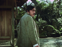 In the simple rock garden of the coastal inn (background), Aochi pauses in the center of the frame and turns back to look at Nakasago and Oine behind him (off-­screen).