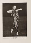 A sepia photograph of Mary Wigman from Death Dance II, here portrayed solo. She wears the chevron-striped draped costume and mask, bending her arms outward and meeting her fingers together below her chin. She stands in a position with one leg behind her and toes pointed atop a carpet floor.