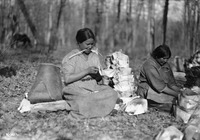 A black-and-white photograph of two women making birch-bark containers on the ground outside.