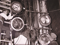 1930s era photograph of two men installing theatrical lighting, one hanging an instrument from a ladder and the other holding a coil of cable.