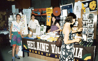 Colorful photo of the HV Press booth packed with displays of postcards, baskets of buttons, and stacks of T-shirts and card sets. Posters, prints, and signs hang behind.