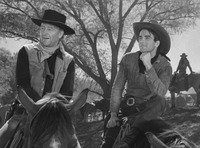The Frontier as Fashion (2): Production still for Red River (1948), featuring Montgomery Clift in a stylized, rather chic cowboy outfit, with impeccable leather chaps and tasselled shirt, as he turns toward a more homely and plainly dressed John Wayne, seated on the Left.