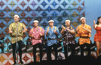 Color photograph of performers (The Drifters) on stage, dancing. Performers wear colorful Japanese festival-­style clothes. Ikariya stands in front of mic singing, while other members dance to his left. On right side of photograph, a woman in a red dress is partially visible.