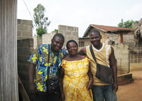 Three smiling people. Two are dressed in African clothing, one in Western clothing. The two young men stand on either side of their mother, each with an arm over her shoulder.