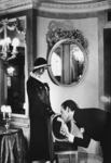 “Upstairs at Maxim’s,” shot by Helmut Newton in the famous Parisian restaurant, depicts a scene of hand-kissing which takes place in a richly-furnished interior – Belle Époque-style mirror, chair, and lighting fixture. An elegant gentleman, wearing a pinstriped suit over a white shirt, bends forward to kiss the extended hand of a stylish woman in a Chanel suit and hat. She casts her gaze down on the man, who places an ardent kiss on her wrist.