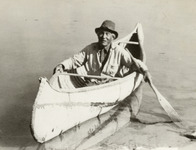 A black-and-white photograph of a figure kneeling in and paddling a canoe.