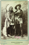 Sitting Bull, wearing a Lakota war bonnet, stands next to Buffalo Bill Cody, who is dressed in his stage costume, including an embroidered satin shirt and tall boots. Cody partly leans on his rifle.