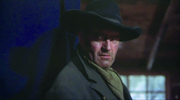 Close-­up from chest to hat of a grizzled John Wayne, his head turned and glaring violently towards the Right of screen. Behind him on the Left, the recessed space of the cabin and a frosty window. On the Right, a blue curtain, its edge bisecting the space. Careful lighting causes the hat brim to cast a dark shadow over his eyes, especially the right, while his body casts a shadow on the curtain as well.