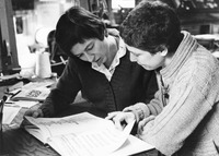 Black-and-white photo. Several books are laid out on a large table in the print shop. Both women look intently at a book opened to typeface examples.