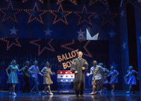 Photograph of the actor playing the Chief Justice sings to the audience from center stage in full judicial robes, with a lit-­up sign that says “Ballot Box” behind him. The ensemble members dance upstage and to the side of him as he sings.