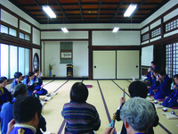 Fig. 14. A photograph of half-circle of people, seated on the floor of a temple room, practicing  with goeika bells in their left hands. Each has a small book in front of them.