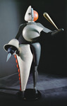 Color photograph of a costume for the Triadic Ballet: an abstracted figure, with clubs and spears for arms and hands, a large white and orange leg that won’t bend, and large circular forms for the torso and head. The head is split: half is white and half is orange.