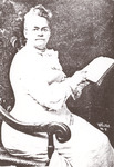 Nation is posed in a chair for a studio portrait wearing a light-colored, long-sleeved dress. She holds a book, probably a Bible, and points to a passage with her forefinger.